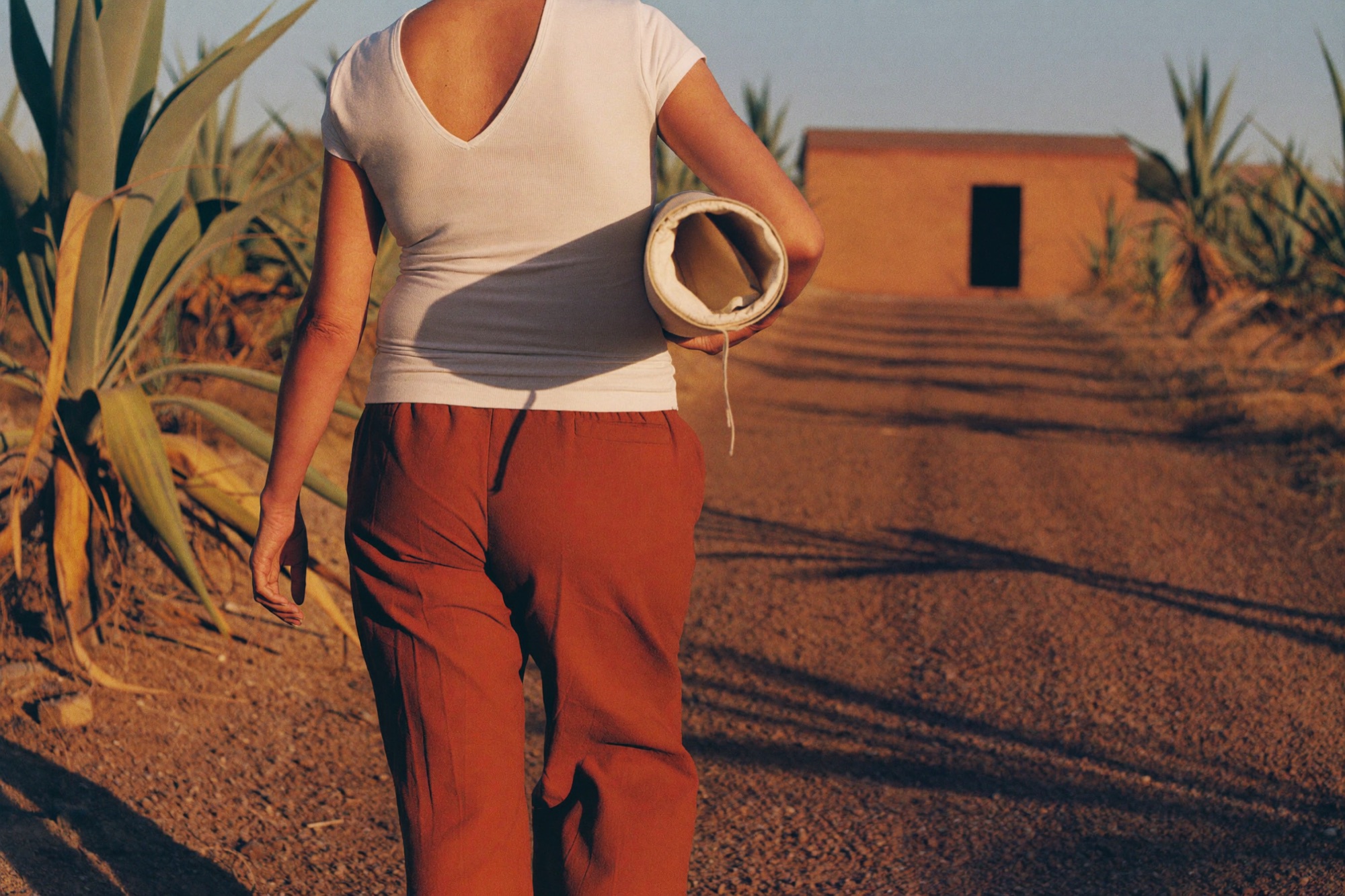 Una mujer con playera de cuello en V camina hacia un taller de adobe cargando un lienzo enrollado